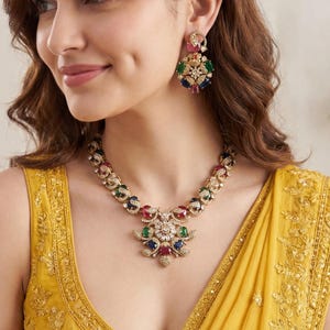 May include: A gold necklace and earring set with colourful gemstones. The necklace has a floral pendant. The model is wearing a yellow top.