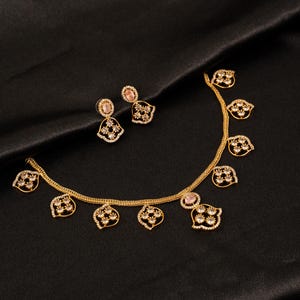 Peut inclure: Un collier doré et une paire de boucles d'oreilles assorties. Le collier présente une chaîne délicate avec des breloques en forme de feuille, chacune ornée de petites pierres claires et d'une pierre ovale rose. Les boucles reprennent ce motif.