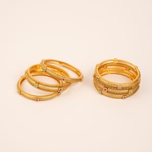 Peut inclure: Un ensemble de bracelets rigides dorés présentant une surface texturée à motifs croisés. Ces bracelets circulaires sont ornés de petites pierres carrées alternant entre le rouge et le vert, incrustées à intervalles réguliers sur la bande.