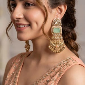 Könnte beinhalten: Eine Frau trägt große, verzierte goldene Ohrringe mit einem rechteckigen grünen Stein und Perlenakzenten. Die Ohrringe haben ein mehrschichtiges Design mit hängenden Goldperlen. Sie trägt ein pfirsichfarbenes Oberteil mit aufwendiger Goldstickerei.