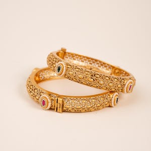 May include: Two gold bangles with detailed, repeating patterns. Each bangle has a clasp and is decorated with small, colourful gemstones, including red, green, and white accents. The jewellery is displayed on a neutral background.