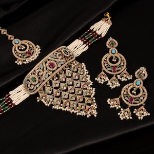 May include: An elaborate gold-coloured jewellery set comprising a necklace, earrings, and a headpiece. The set is adorned with colourful gemstones, pearls, and intricate patterns. The jewellery is presented against a black backdrop.