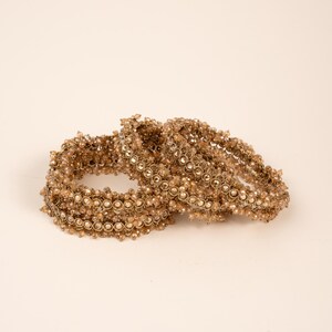 May include: A collection of gold-coloured bangles, set against a neutral backdrop. The bangles are adorned with small, round beads and decorative features, resulting in a textured and elaborate look. The jewellery appears to be crafted from metal and beads.