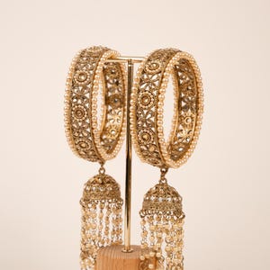 Peut inclure: Deux bracelets dorés ornés de perles et de pendentifs en forme de cloche. Les bracelets sont exposés sur un présentoir doré avec une base en bois. Les bijoux présentent des détails complexes et une esthétique vintage.