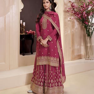 May include: A magenta salwar kameez outfit with gold embroidery. The long-sleeved top and wide-legged trousers are adorned with detailed gold patterns. A matching scarf with gold edging completes the ensemble. The woman is wearing a headpiece and earrings.