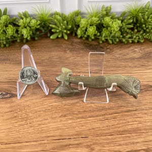 May include: A small, olive-green stone axe displayed on a clear acrylic stand. The axe features a detailed carved design and is positioned next to a quarter coin, also on a stand. The background includes green plants and a white wall.