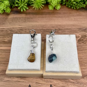 May include: Two silver-toned metal keychains featuring polished tumbled stones. Keychain A has a brown and gold striped stone, while keychain B features a dark blue and green stone. Both include a silver lobster clasp and a circular ring.