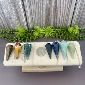 May include: A collection of gemstone pendulums in various colors, including brown, yellow, green, blue, and teal, displayed on a white rectangular stand. Each pendulum is suspended from a silver chain. A US quarter is placed next to the pendulums for size comparison.