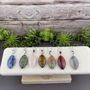 May include: A collection of seven gemstone pendant necklaces displayed on a white stand. The gemstones are oval-shaped and vary in color, including blue, green, pink, brown, red, and gray. Each necklace has a silver-colored chain.