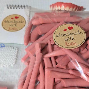 Puede incluir: Se muestran palitos de cera dental rosa y un juego de dentaduras postizas. Se ve un pequeño cuaderno con una pegatina "Handmade with love". También hay una bolsa de pequeñas cuentas blancas.