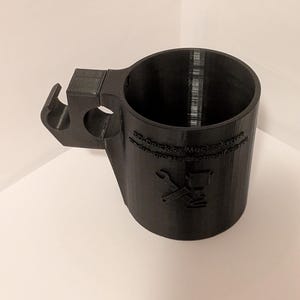 Puede incluir: Taza negra impresa en 3D con asa y diseño único. La taza presenta un cuerpo cilíndrico con un asa que tiene una extensión en forma de gancho. La taza tiene texto y un logotipo en el lateral. La taza está hecha de material negro mate.