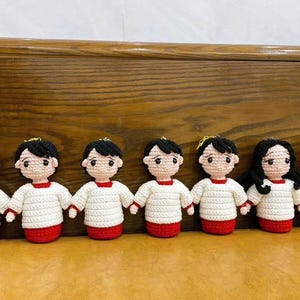 May include: A row of seven handmade crochet dolls. Each doll has black hair, a white shirt with red trim, and a red base. The dolls are holding hands, creating a unified display. The dolls are approximately 4 inches tall.