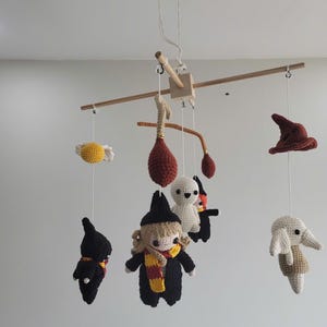 Può includere: Mobile per neonati fatto a mano all'uncinetto, con vari personaggi e oggetti. Il mobile include un boccino d'oro, un cappello parlante e figure in abiti neri con sciarpe. Sospeso a un telaio di legno.