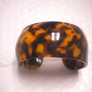 May include: A wide, open-cuff bracelet with a tortoiseshell pattern. The bracelet features a mix of dark brown and amber tones, creating a marbled effect. The cuff is a solid, curved shape, suitable for wrist wear.