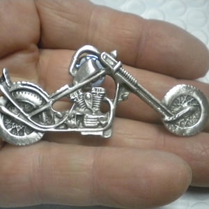 Puede incluir: Un broche de metal plateado con forma de motocicleta chopper. El diseño presenta una horquilla delantera larga, un bloque de motor detallado y ruedas de radios. La superficie tiene un acabado mate texturizado con detalles metálicos en relieve.