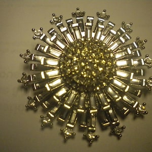 May include: A silver-toned brooch with a sunburst design. The center is a cluster of small, round, gold-colored stones. Elongated, rectangular silver pieces radiate outwards, interspersed with small, star-shaped accents.