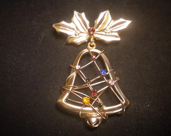 May include: Gold-toned Christmas bell brooch with colorful rhinestones and a holly leaf accent. The bell is adorned with red, blue, yellow, and clear rhinestones, and the holly leaf has a red rhinestone center.