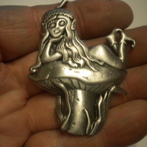 May include: A silver-toned metal pendant featuring a figure with long hair and a cap resting on top of a large mushroom. The figure is depicted in a reclining position with one hand supporting the head, and the pendant includes a small loop at the top.