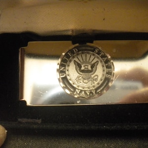 May include: A silver-toned money clip with a circular emblem featuring the words "United States Navy" and an eagle with arrows and an anchor. The clip is rectangular and sits in a black velvet-lined box.
