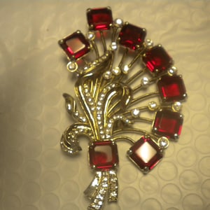 May include: A gold-coloured brooch in a floral design, with square, deep red gemstones and clear, round rhinestones. The brooch has a ribbon-like detail at the base, set against a textured background.