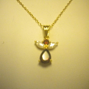 May include: A gold-toned necklace with a pendant shaped like an angel. The pendant features a teardrop-shaped, dark red gemstone, a small round red gemstone, and two white, wing-shaped gemstones. The chain is a delicate, linked design.