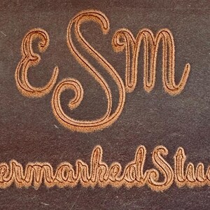 Könnte beinhalten: Nahaufnahme einer dunkelbraunen Oberfläche mit den Worten „eSm“ und „Evermarked Studio“ in einer dekorativen, hellbraunen Schriftart. Die Buchstaben sind stilisiert mit einem strukturierten, geschnitzten Aussehen, was auf ein Logo oder ein Branding-Element hindeutet.