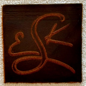 以下が含まれることがあります： 彫刻された、様式化された文字「S」と装飾的な要素が特徴の、正方形の濃い茶色の革製コースター。コースターの端は明るい茶色です。デザインは暖かく赤みがかった茶色です。
