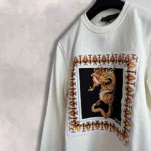 May include: Cream-coloured sweatshirt with a gold and orange dragon design on a black background, framed by an ornate border. The sweatshirt has a crew neck and long sleeves. The brand name is visible on the label.