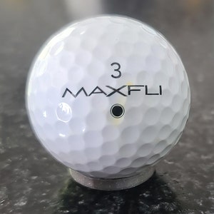May include: A white golf ball with the text "MAXFLI" and the number "3" printed on it. The ball has a black dot in the center.