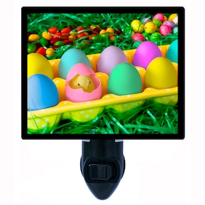 May include: A yellow egg carton filled with colorful Easter eggs, including a pink egg with a baby chick peeking out. The carton is surrounded by green grass and candy.