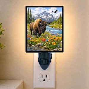 May include: A rectangular plug-in night light featuring a vibrant landscape scene. The image displays a brown bison standing in a field of colorful wildflowers near a flowing river, with snow-capped mountains and a flying bird in the background.