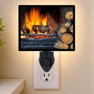 May include: A rectangular plug-in night light featuring a printed image of a burning fireplace with bright orange flames and a stack of brown logs on the right side. The light has a black frame and a black base with a switch, plugged into a wall outlet.
