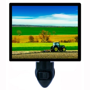 May include: A green tractor plows a field with a blue sky and white clouds in the background. The field is a mix of green and brown.