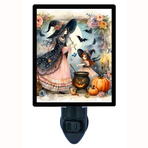 May include: A nightlight featuring a whimsical illustration of two witches in front of a cauldron with a jack-o'-lantern face. The witches are wearing black hats and pink dresses with stars and floral accents. The background includes a full moon, bats, and candles.