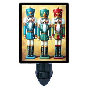 May include: A decorative night light featuring a framed image of three colorful nutcracker soldiers. Each nutcracker wears a different colored uniform: blue, green, and red. The night light has a black border and a black plug.