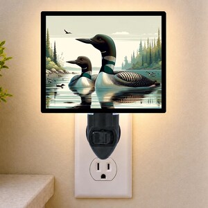May include: A rectangular plug-in night light featuring a graphic illustration of two loons swimming on a calm lake. The scene includes a forest shoreline, a soft sky, and a small bird in flight. The light is plugged into a standard wall outlet.