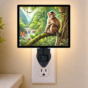 May include: A rectangular plug-in night light featuring a black frame and a vibrant jungle scene. The image displays a brown monkey perched on a mossy tree branch, surrounded by lush green foliage, tropical plants, and a distant waterfall.