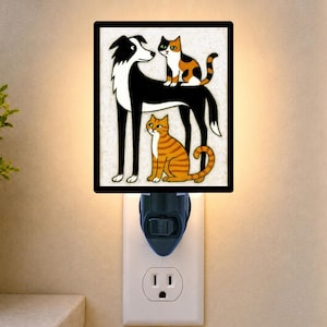 May include: A square plug-in night light featuring a black and white dog standing with a calico cat perched on its back and an orange tabby cat sitting between its legs. The illustration is set against a textured off-white background with a black border.