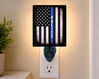 May include: A rectangular plug-in night light featuring a distressed American flag design with a vertical blue stripe and the word POLICE printed in white capital letters. The light emits a warm glow from behind the black-bordered graphic panel.