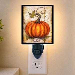 May include: A rectangular plug-in night light featuring a vibrant orange pumpkin illustration on a weathered, light-colored wood-grain background. The design includes green leaves, a dark stem, and delicate vine tendrils, all framed by a black border.