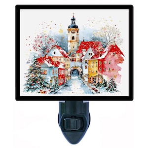 May include: A night light featuring a watercolor painting of a European village scene with red-roofed buildings, a church tower, and snow-covered streets. The artwork is framed in black and set against a white background.