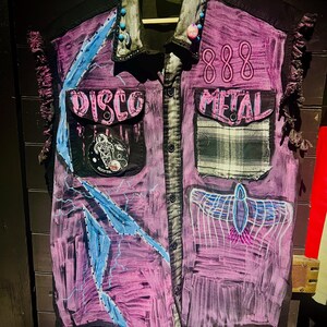 May include: A sleeveless, button-down shirt in shades of purple, black, and blue, adorned with patches and text. The shirt features the words "DISCO" and "METAL", along with the number "888". Decorative beads are attached to the collar.