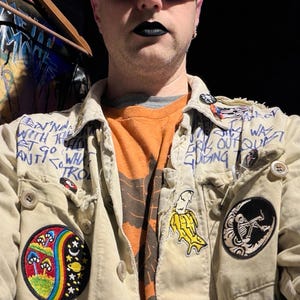 May include: A person wearing rainbow-tinted sunglasses and black lipstick. They are wearing a tan jacket with patches, including a mushroom design, a banana, and a skeleton. The jacket has handwritten text.