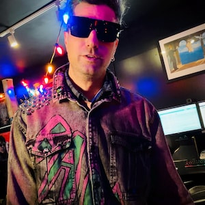 May include: A denim jacket with a pink and green design, adorned with silver studs on the shoulders. The jacket features a button-down front and two chest pockets. The person in the image is wearing black sunglasses.