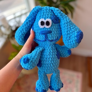May include: A handmade, plush blue dog with dark blue spots and accents. The dog has large, floppy ears, white and black eyes, and a stitched smile. The toy is made of a soft, textured material, perfect for children.