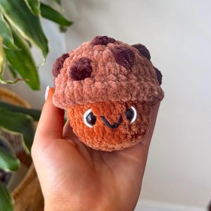 May include: A small, round, crocheted plush toy shaped like a muffin or cupcake. The top is a light brown with dark brown spots, while the base is a solid orange-brown. It features two black plastic eyes with white outlines and a stitched black mouth.