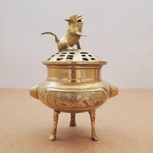 Peut inclure: Un brûleur d'encens doré avec une figurine de lion décorative sur le dessus. Le brûleur a un corps rond avec des motifs de feuilles et trois pieds. Le couvercle est perforé de trous rectangulaires.