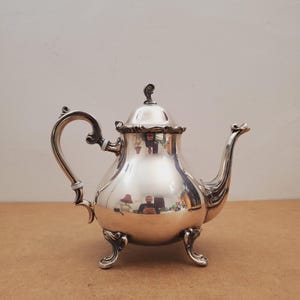 Op de afbeelding: Een zilveren theepot met een gebogen handvat en tuit, rustend op drie sierlijke voetjes. De theepot heeft een reflecterend oppervlak en een kleine decoratieve knop op het deksel. Het ontwerp is klassiek en elegant.