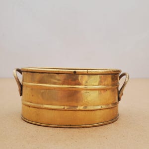 May include: A small, round, polished brass basin featuring two curved side handles. The container has a smooth, metallic surface with two horizontal ridges encircling the body, providing a simple, functional design for holding small items.