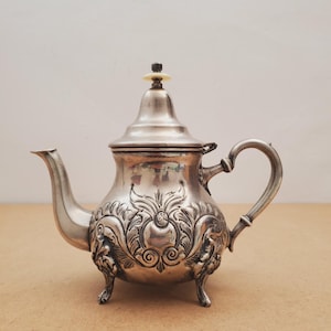 May include: A silver-colored teapot with ornate floral and scrollwork designs. The teapot has a curved spout, a heart-shaped handle, and three small feet. The lid is topped with a small, light-colored knob.